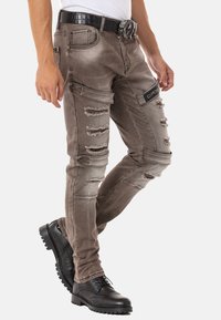 Braune, zerrissene Jeans aus strukturiertem Stoff, mit Seitentaschen und einem Logo-Patch. Kombiniert mit einem schwarzen Gürtel mit Krokodilembossierung und schwarzen Schuhen.