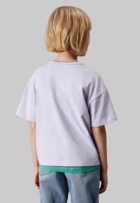 Camiseta de algodón color lavanda claro con un corte relajado, mangas cortas y cuello redondo, superpuesta sobre una camisa verde, combinada con jeans de color azul claro.