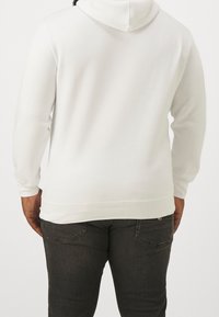 Persona che indossa una felpa bianca semplice e jeans grigio scuro, mostrata di spalle su uno sfondo chiaro e uniforme.