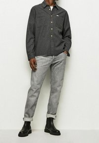 Camisa gris abotonada con dos bolsillos en el pecho, combinada con jeans gris claro y botines negros. La camisa tiene un cuello con solapa y botones de madera.