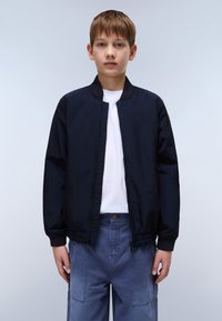 Η ναυτική μπλε τζάκετ bomber με φερμουάρ μπροστά, ριμπ κολάρο και ελαστική ζώνη μέσης. Συνδυασμένο με ανοιχτό μπλε παντελόνι στυλ cargo.