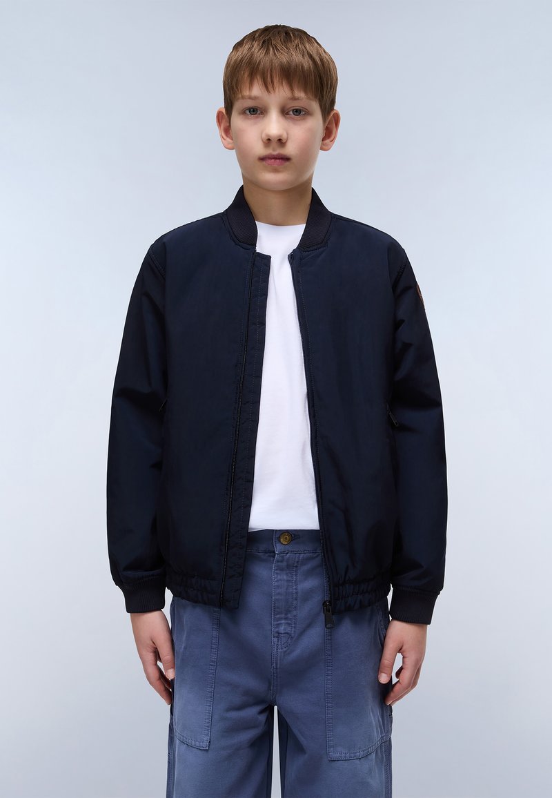 Η ναυτική μπλε τζάκετ bomber με φερμουάρ μπροστά, ριμπ κολάρο και ελαστική ζώνη μέσης. Συνδυασμένο με ανοιχτό μπλε παντελόνι στυλ cargo.