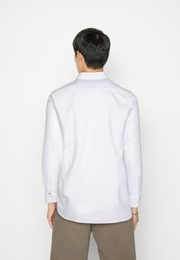 Camisa blanca de manga larga con cuello abotonado, tejido texturizado y dobladillo redondeado; presenta puños abotonados y un pequeño detalle de logo.