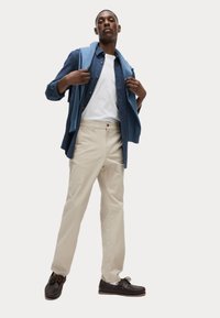Beige Hosen, weißes T-Shirt, dunkelblause Hemd mit Knopfleiste und hellblauer Pullover über die Schultern geschlungen. Braune geschnürte Schuhe.