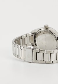 Montre en acier inoxydable avec un bracelet poli, un boîtier rond et un cadran argenté. Présente un logo gravé sur le fermoir et un fond texturé.