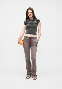 Νεαρή γυναίκα που φοράει στενό σκούρο γκρι κορμάκι και φαρδιά ξεθωριασμένα καφέ παντελόνια, κρατώντας μια μικρή πορτοκαλί τσάντα και φορώντας μαύρα σανδάλια.