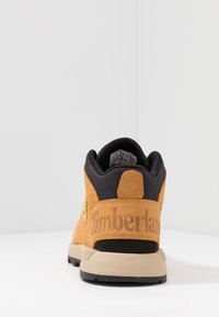 Botine de gleznă din suede bej, cu guler căptușit negru, talpă intermediară bej, talpă exterioară neagră și branding embosat "Timberland" pe călcâi.