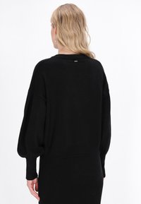 Czarny, oversize sweter z długimi, bufiastymi rękawami i ściągaczem u dołu. Miękka struktura, prosty design, z małym metalowym akcentem na plecach.
