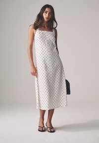 White midi dress with black polka dots, square neckline, thin straps, and a straight silhouette. Paired with black flip-flops and bag.