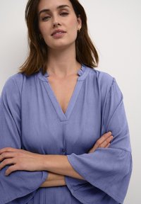 Svetlofialová, textúrovaná blúzka s výstrihom do V, riaseným golierom a širokými zvonovými rukávmi. Model pózuje so zlomenými rukami proti neutrálnemu pozadiu.