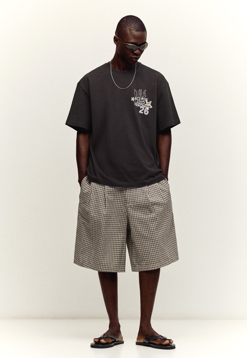 Homme portant un t-shirt noir graphique, un short à carreaux surdimensionné, des sandales noires, des lunettes de soleil et une chaîne argentée, debout les mains dans les poches.