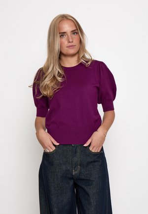 Giovane donna con lunghi capelli biondi che indossa un maglione viola a maniche corte e jeans blu scuro a gamba larga con le mani nelle tasche.