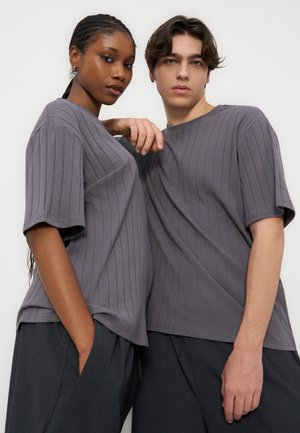 Δύο μοντέλα φορούν ταιριαστά γκρι ριγωτά κοντομάνικα μπλουζάκια και σκοτεινό παντελόνι, στέκονται κοντά με ουδέτερες εκφράσεις μπροστά σε λευκό φόντο.