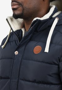 Tmavomodrá vatovaná bunda s kožušinovým golierom, zipsovým zapínaním, krémovými sťahovacími šnúrkami a hnedou koženou nášivkou s logom na hrudi.