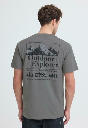 Man die wegkijkt, draagt een grijs t-shirt met een bergafbeelding en de tekst "Outdoor Explorer" en bomen aan de achterkant.