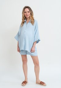 Chemise légère bleu clair, ample, à manches courtes, avec des boutons, assortie à un short; tissu texturé, détail de boutonnage simple, et sandales beiges.