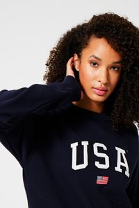 Navy sweatshirt with white "USA" lettering and small flag detail. Features a round neckline and ribbed cuffs. Fabric appears soft and textured.