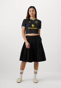 Camiseta curta preta de manga curta com detalhes em amarelo e azul, combinada com uma saia plissada preta. Meias brancas com riscas castanhas e sapatos estampados.