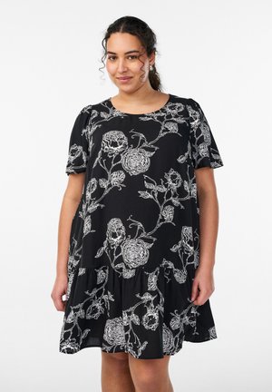 Frau in einem schwarzen, kurzärmeligen Kleid mit weißen Blumenmustern steht vor einem schlichten weißen Hintergrund.