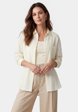 Donna con lunghi capelli castani che indossa una camicia crema con bottoni sopra una maglietta beige e pantaloni beige, in piedi davanti a uno sfondo bianco.