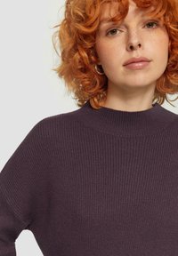 Lila gerippter Pullover mit hohem Hals und lockerer Passform, der eine weiche Textur und subtile Details in der Stoffverarbeitung zeigt.