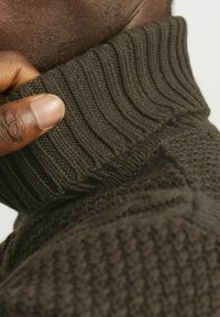 Primo piano di un maglione di lana marrone, con colletto a coste, trama testurizzata e una mano visibile che tiene il colletto.