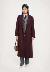 Burgunderfarbener Wollmantel mit breiten Revers, doppelreihigem Design, kombiniert mit einer karierten Weste und maßgeschneiderten grauen Hosen. Weiße spitze Schuhe und braune Tasche.