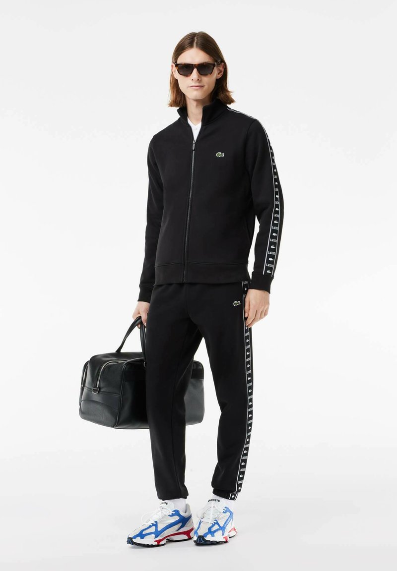 Junge Person trägt eine schwarze Zip-Jacke und Hose mit Logo-Streifen, weiße Sneaker mit blauen und roten Akzenten, Sonnenbrille und hält eine schwarze Tasche.