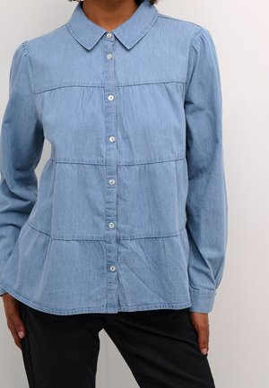 Kvinde iført en lyseblå langærmet skjorte med knapper, lagdelte syninger og Peter Pan-krave, kombineret med sorte bukser.