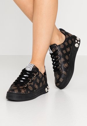 Baskets noires avec motif de logo marron et gros strass au talon, portées pieds nus sur un fond clair uni.