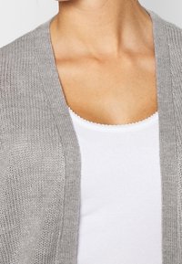 Cardigan a maglia grigia chiaro con apertura frontale, caratterizzato da una texture a coste, accompagnato da un top attillato bianco con scollatura a onda.