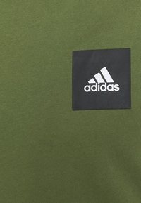 Oliwkowa koszulka z gładką fakturą, z kwadratową czarną naszywką, na której widnieje białe logo Adidas. Prosty dekolt typu crew.