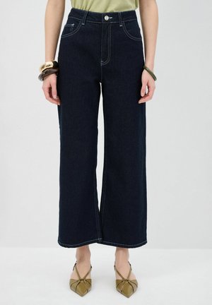Jeans de mezclilla oscura de pierna ancha con cintura alta, costuras blancas en contraste, dos bolsillos frontales y cierre de botón. Combinados con calzado verde oliva.
