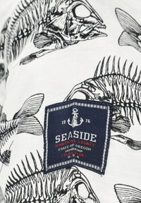 Biały materiał z czarnymi nadrukami szkieletu ryby i granatową naszywką z napisem "Seaside Nautical Legacy State of Oregon Membership SSNL Club USA 1976."