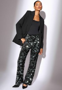 Črn blazer preko prilegajoče se črne majice; sijoče črne hlače s sequini z teksturiranim vzorcem; ostri črni salonarji; model samozavestno pozira.