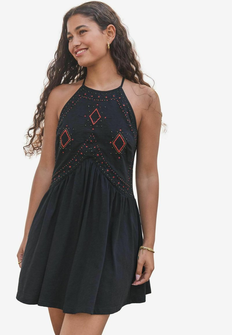 Svart ärmlös klänning med röda och vita broderade diamantmönster, en figurnära bodice och en utsvängd kjol. Slät bomullstextur.