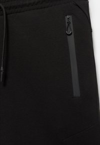 Gros plan d’un pantalon en tissu noir montrant une ceinture élastique, un cordon de serrage et une poche verticale zippée sur le côté droit.