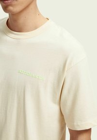 T-shirt color crema realizzato in morbido cotone, con scollatura rotonda e un piccolo logo verde chiaro sul petto. Maniche corte, vestibilità comoda.