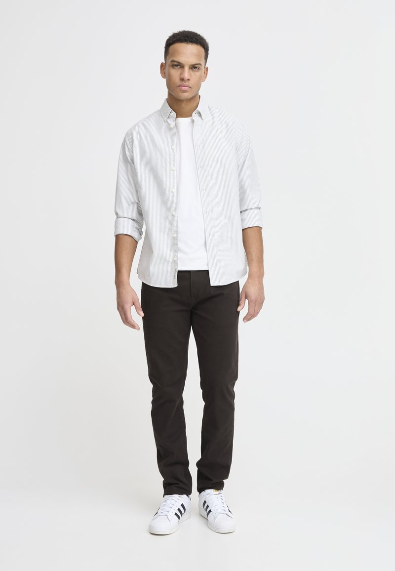 Chemise à boutons avec de fines rayures verticales, portée par-dessus un T-shirt blanc uni. Assortie avec un pantalon sombre et des baskets blanches avec des rayures noires.