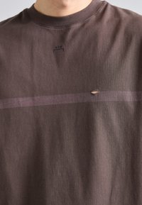 Καφέ βαμβακερή μπλούζα κοντομάνικη με στρογγυλή λαιμόκοψη, με οριζόντια μωβ ρίγα και μια μικρή τρύπα κοντά στο κέντρο. Λογότυπο τυπωμένο πάνω από τη ρίγα.