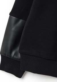 Mangas de sudadera negra con puños de canalé, una manga presenta un detalle de parche de cuero negro liso.