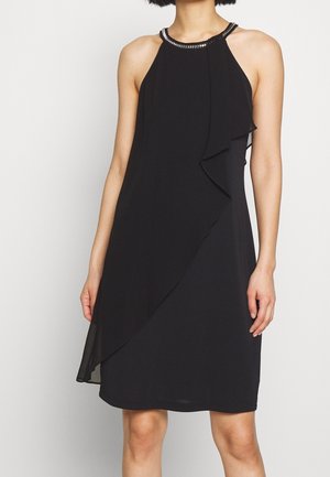 Frau trägt ein ärmelloses schwarzes knielanges Kleid mit Perlenbesetztem Neckholder-Ausschnitt und asymmetrischem Rüschendetail auf der rechten Vorderseite.