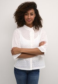 Blusa bianca in pizzo con maniche a sbuffo corte, scollo rotondo e chiusura a bottoni. Presenta intricate decorazioni a taglio. Indossata sopra dei jeans blu.