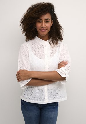 Blusa bianca in pizzo con maniche a sbuffo corte, scollo rotondo e chiusura a bottoni. Presenta intricate decorazioni a taglio. Indossata sopra dei jeans blu.