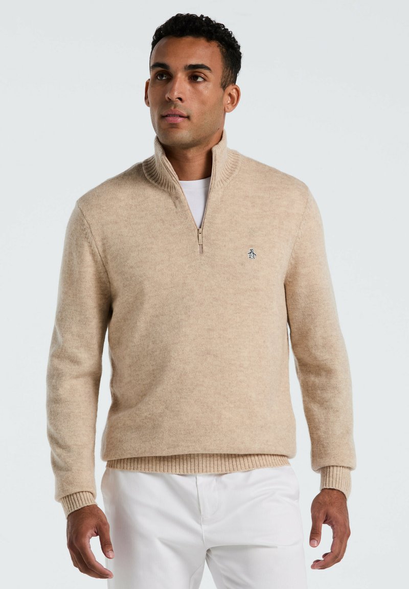 Beige Strickpullover mit Reißverschluss aus weichem Material. Gerippte Bündchen und Saum, mit einem kleinen Logo auf der Brust. Kombiniert mit weißen Hosen.