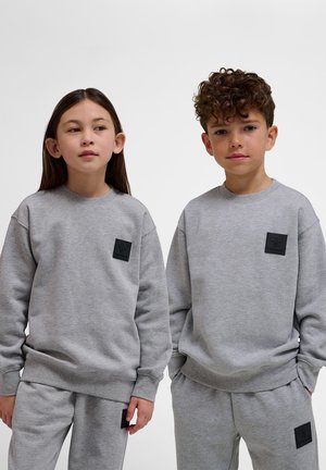 Dwoje dzieci stojących obok siebie w pasujących szarych bluzach i spodniach dresowych z czarnymi naszywkami z logo, neutralne wyrazy twarzy, jednolite tło.