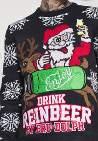 Pull en tricot avec un motif coloré de Père Noël et de renne, accompagné d'une canette verte étiquetée "Enjoy". Des motifs de flocons de neige décorent le fond sombre.