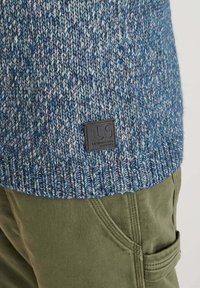 Stickad tröja i blå och grå mélange, med en räfflad nederkant och en svart märkespatch. Bärs över olivgröna byxor med sidfickor.