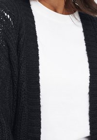 Svart stickad cardigan med ett strukturerat mönster, öppen front och ribbade kanter, bärs över en enkel vit t-shirt. Mjuk stickad kvalitet.