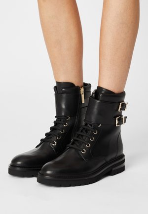 Czarne skórzane botki za kostkę z sznurowadłami, bocznymi zamkami błyskawicznymi i podwójnymi złotymi klamrami noszone na nagich nogach na jednolitym tle.